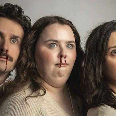 Three actors wearing beige clothes lined up together, noses bleeding and looking startled. 