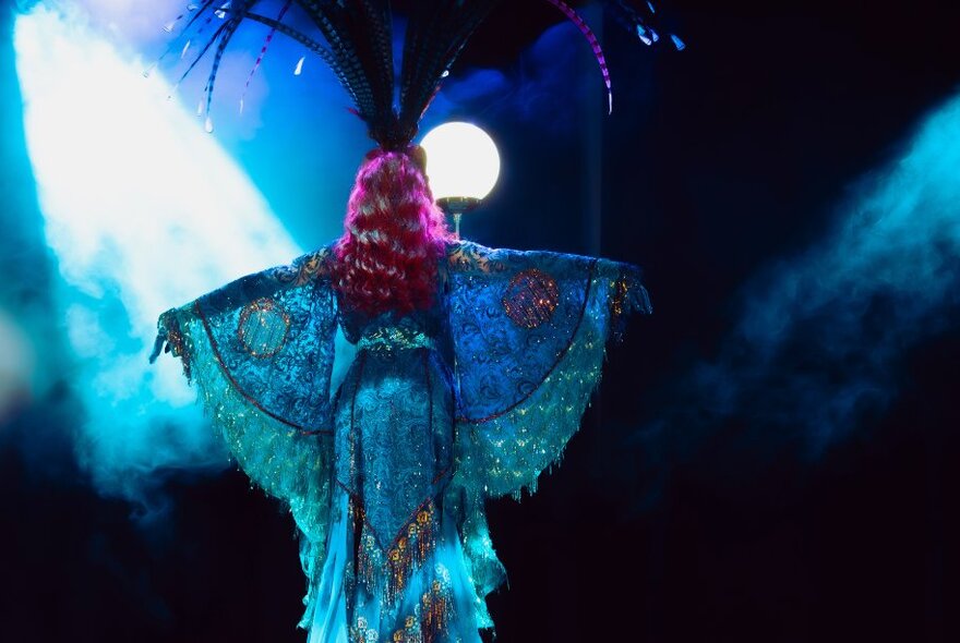 Woman wearing huge feathered headdress standing under lights with arms outstretched.