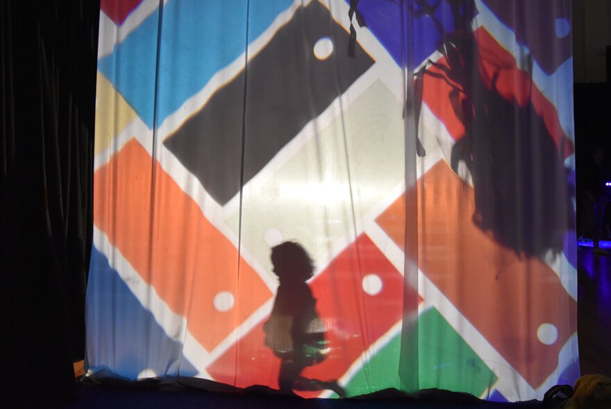 A child's silhouette cast against a brightly patterned geometric backdrop with hanging fabric and shadow play, captured during an interactive family creative program at ArtPlay.