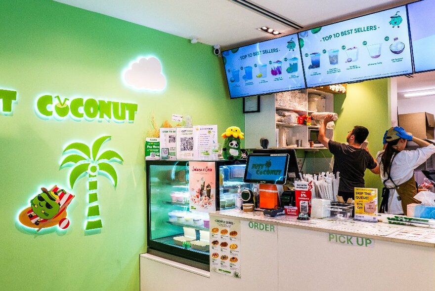 The green walled interior of the cafe Just Coconut  with the name of the shop and a palm tree design painted on the left side wall, and two staff working behind the small service counter, a neon menu hanging near the ceiling.