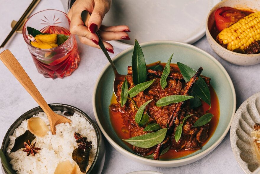 A hand holding a spoon in a bowl of curry, with rice in a side bowl and a red-coloured cocktail in a short tumbler, all on a white table.