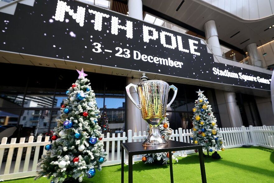 The AFL premiership cup in front of Marvel Stadium and two Christmas trees.