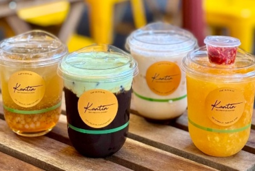 Assorted drinks and bubble teas served in clear plastic takeaway cups, all with a yellow Kantin logo sticker, lined up on a wooden table.