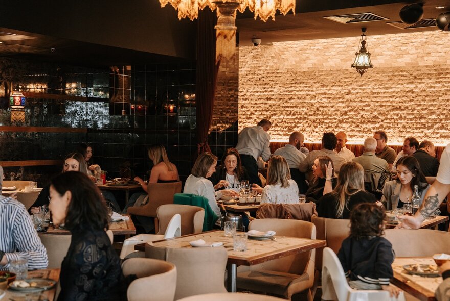 Diners seated at tables at Byblos restaurant, with an exposed light coloured brick wall in the background, and a bar area to the right of frame.