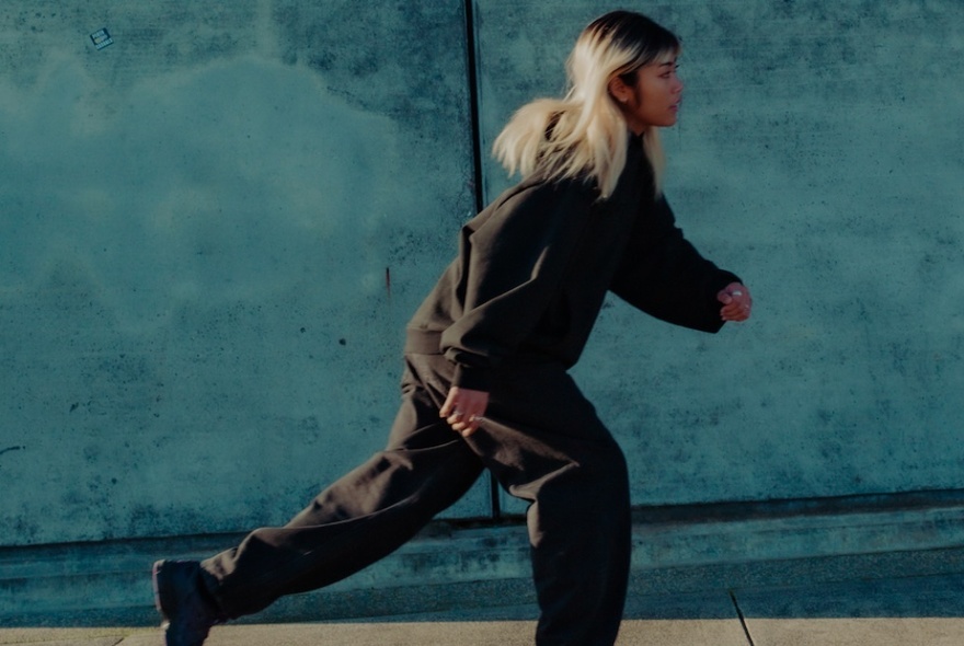 A woman wearing a dark green hooded jumper and tracksuit pants, captured mid-stride in front of a blueish concrete wall.