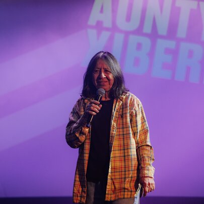 Comedian Aunty Annette doing stand-up in front of a purple backdrop with her name projected onto it. 