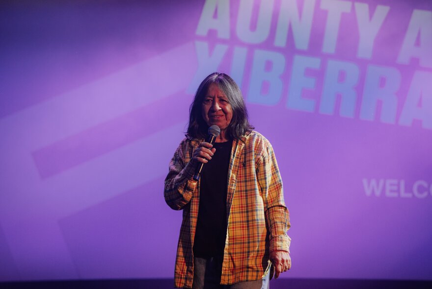 Comedian Aunty Annette doing stand-up in front of a purple backdrop with her name projected onto it. 