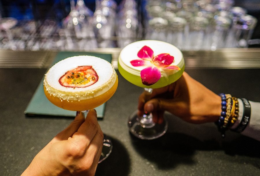 Two people holding colourful foamy cocktails with a flower and passionfruit garnish.