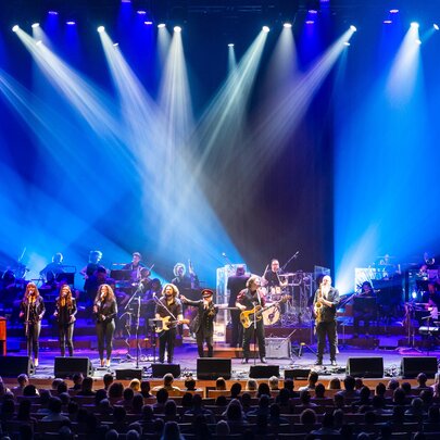 Large rock ensemble performing on stage under blue lights with seated audience.