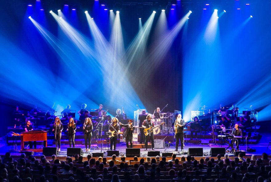 Large rock ensemble performing on stage under blue lights with seated audience.