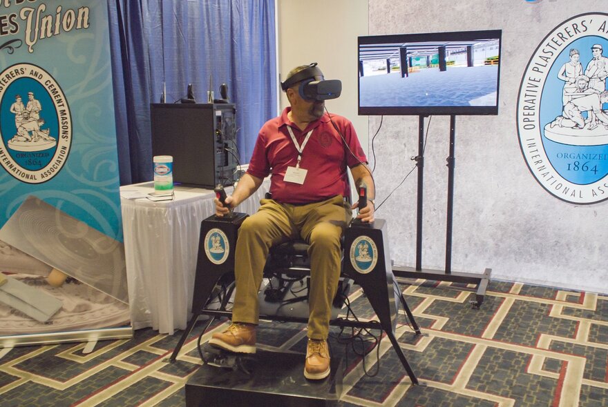 A still from the documentary film, The History of Concrete, shows a man wearing a VR headset sitting in a black mechanical simulator chair, holding joysticks, at a trade show booth with banners and a display monitor in the background.