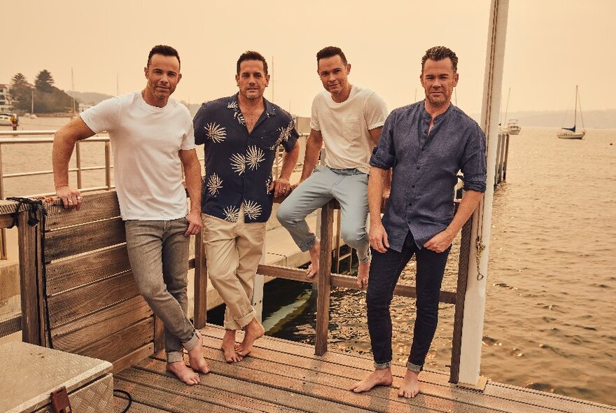 Four men wearing casual summer trousers and shirts, seated and standing on a wooden deck overlooking a yachting harbour, wearing bare feet.