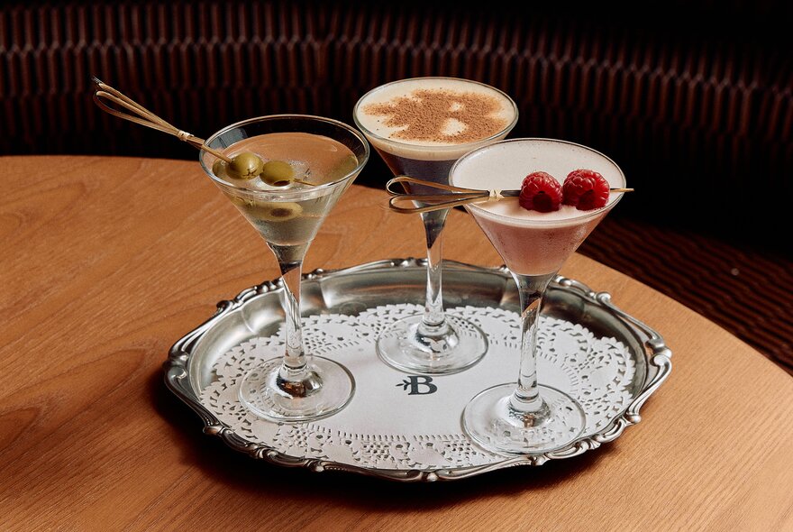 Three different cocktails in martini glasses on a silver platter with a white doily.