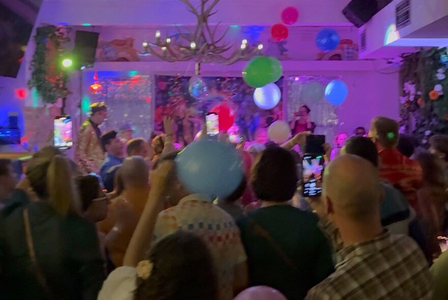A room of people dancing and enjoying themselves while a band plays live on a small stage, balloons floating close to the ceiling.