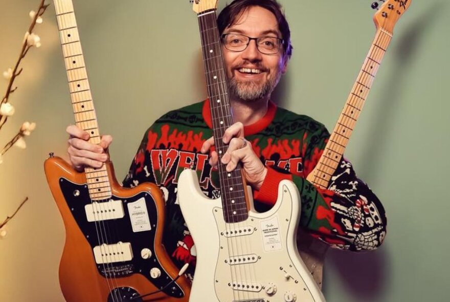 A happy guy with a beard and glasses holding three electric guitars. 