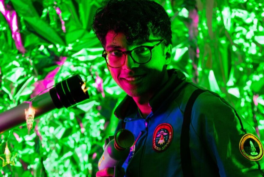Smiling man wearing glasses in half shadows, with a telescope next to him, standing in front of lush greenery.