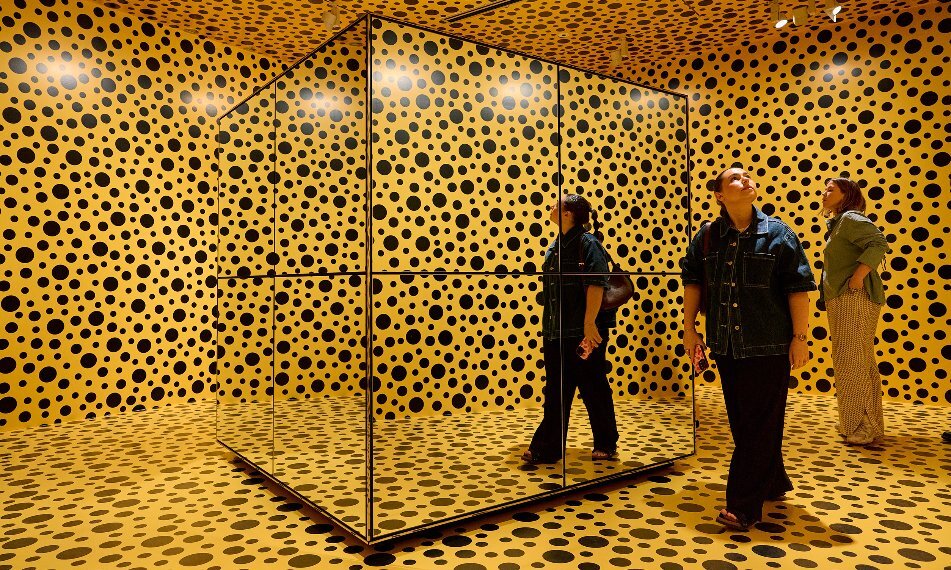 Two women are in a yellow room in a gallery covered in black spots.