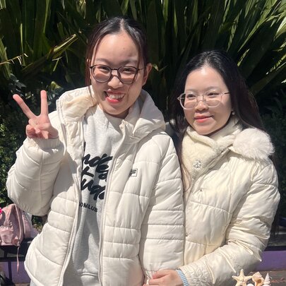 Two individuals wearing matching white quilted puffer jackets at an outdoor market in front of green foliage, with one person making a peace sign.