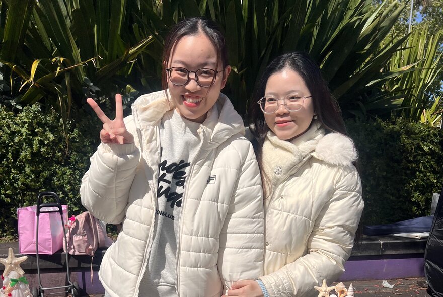 Two individuals wearing matching white quilted puffer jackets at an outdoor market in front of green foliage, with one person making a peace sign.