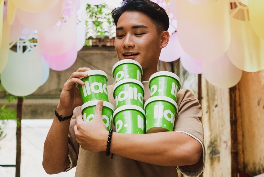 Man with short dark hair holding multiple lime green yoghurt tubs, pastel-white lights above.