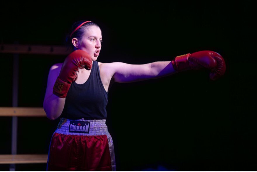 A scene from the play Bitch Boxer, featuring a performer in red boxing shorts and a black singlet top shadow boxing on a darkened stage.