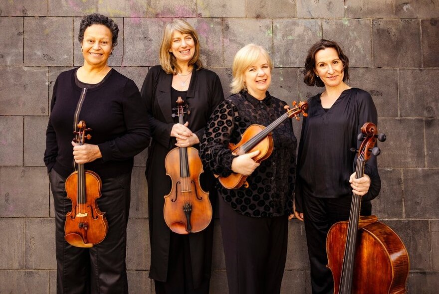 Four female classical musicians holding stringed instruments, wearing black, standing against a grey brick wall.