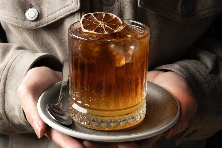 Hands holding a tea-coloured beverage in a glass, garnished with a slice of dried lime. 