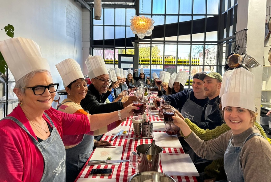 A long table with a red and white checked tablecloth and people seated wearing tall chef's hats, toasting with drinks raised and smiling. 