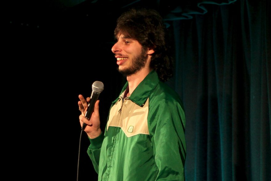 Comedian and mime artist Con Coutis holding a microphone and performing stand-up on a stage.