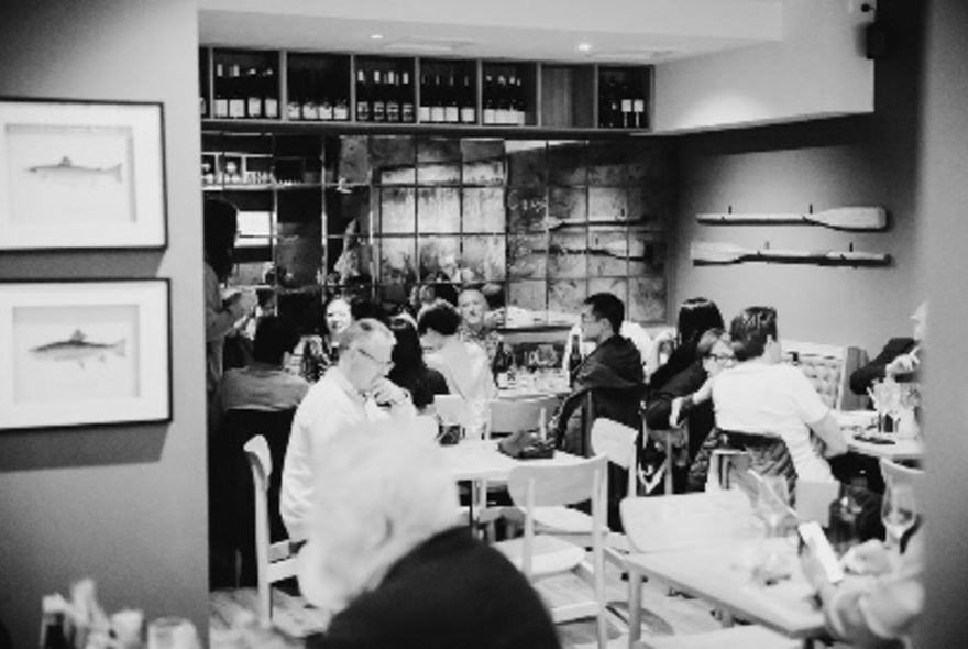 Interior of small wine bar with people dining at tables, framed prints on the wall and a high wall shelf lined with bottles of wine; black and white.