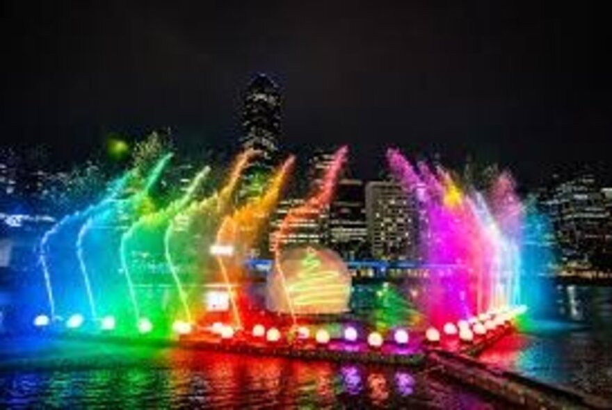 Bright lights and water fountains near the edge of the Yarra River, with the city skyline in the background, as seen from a boat cruise at night.