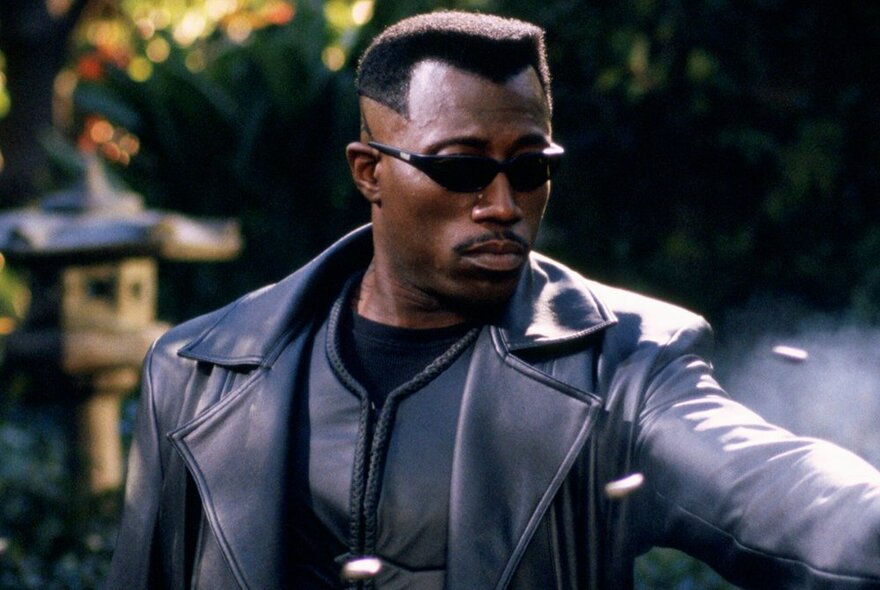 A still from the movie Blade showing the actor dressed in black leather, wearing dark sunglasses and with his arm stretched out in front of him.