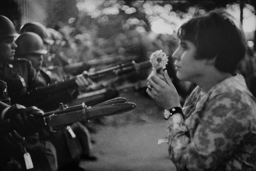 A woman holding a flower standing in front of a row of armed soldiers.