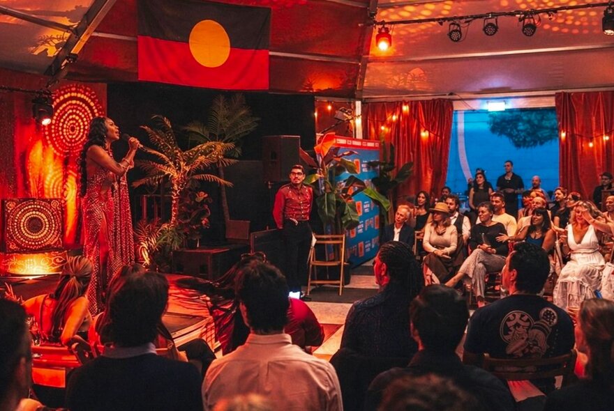 The interior of the Spiegel Haus with a First Nations performer on the stage in front of a crowd, the Aboriginal Flag hanging from the ceiling.