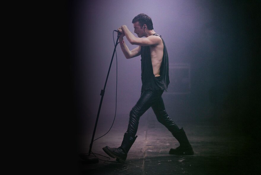 A person in profile on a stage, wearing black tight pants, a black singlet and black boots holding a microphone stand as if about to sing into the microphone.
