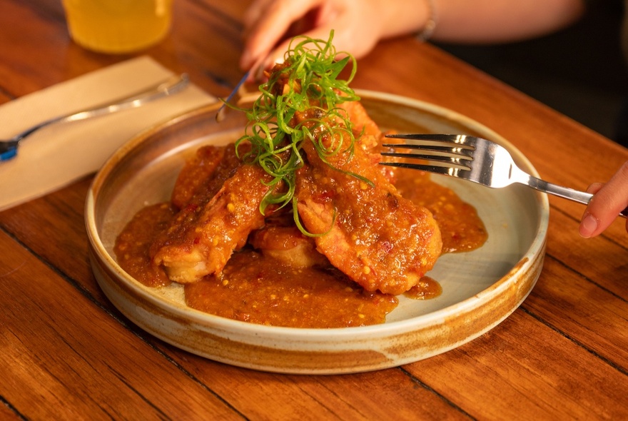 An Asian chicken dish presented in an earthenware dish on a wooden table, a person's hands holding cutlery as if to begin eating.