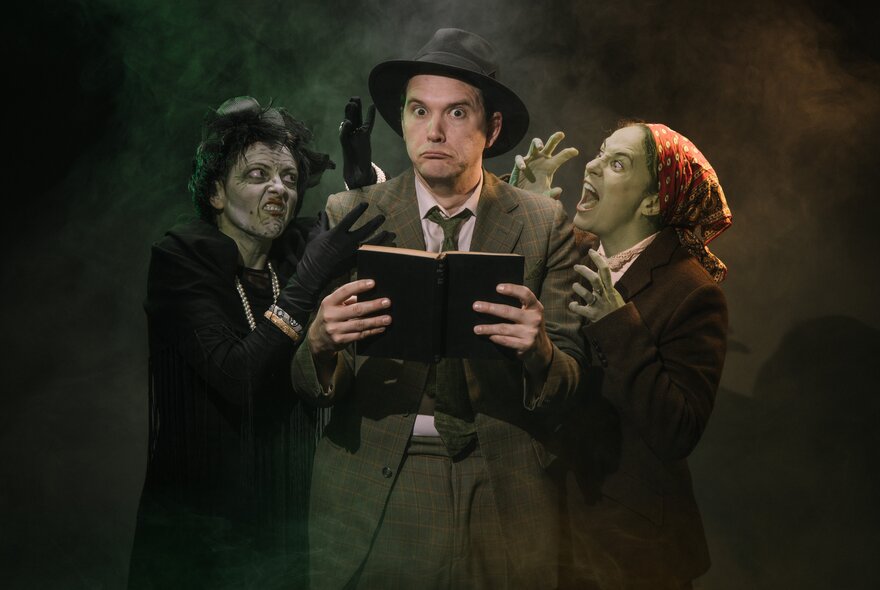 Three actors of an impro ensemble, two dressed as greenish coloured ghouls, and the central character wearing a suit and hat, holding an open book.
