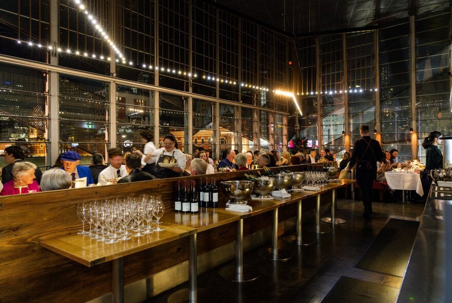 Long bar area with rows of glasses and bottles, dining patrons seated behind with large windows and city views.