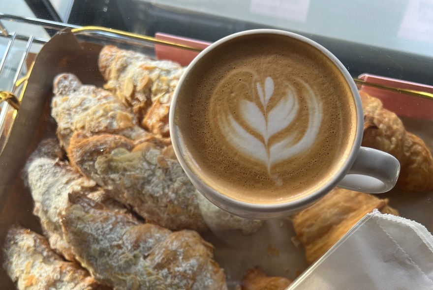 A latte in a white mug with a flower pattern in the foam, resting on a tray of almond croissants.