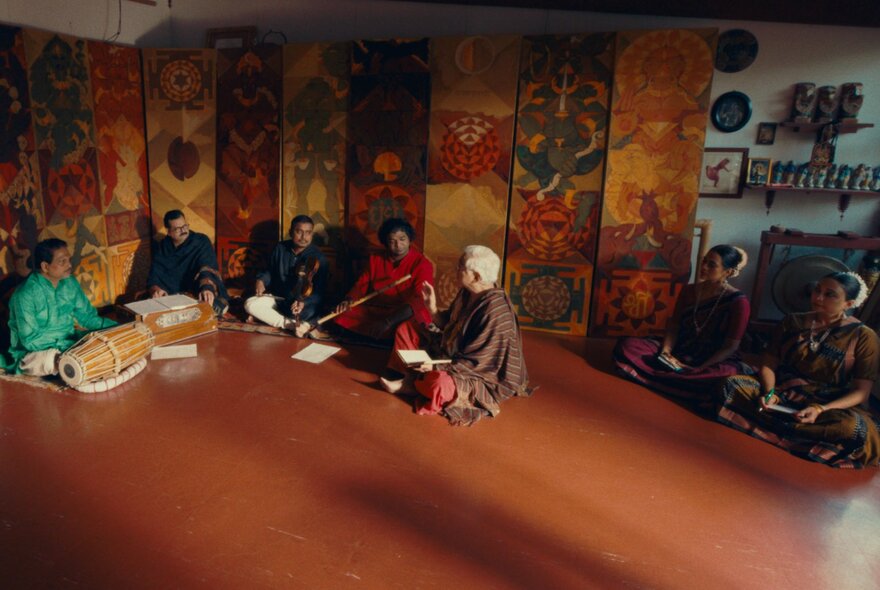A group of Indian musicians sitting cross-legged on the floor with their instruments, colourful Indian motifs on the wall behind them.