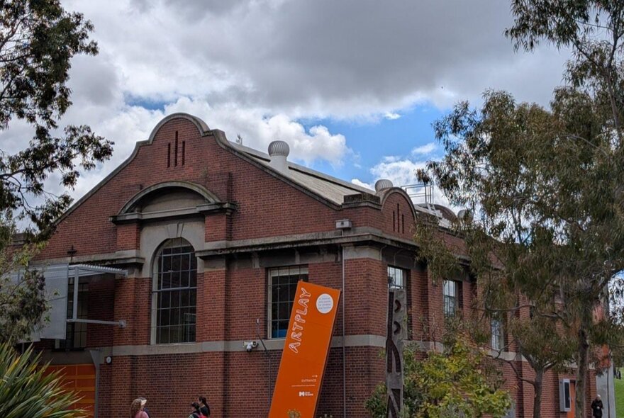 The red brick exterior of the ArtPlay building in Birrarung Marr, with trees on either side of it.