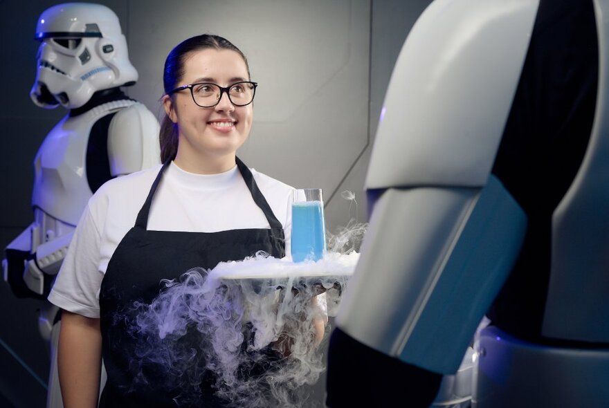 A smiling waiter holding a tray with a glass of blue milk on it, dry ice smoke falling off the edges of the tray, a Star Wars Stormtrooper character in the background. 