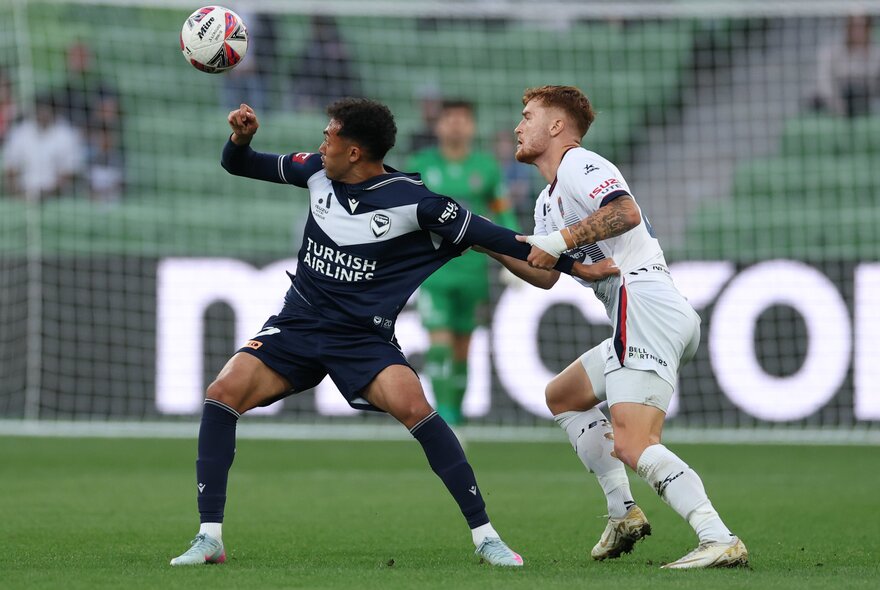 Two soccer players tussle over a ball which is in the air at a soccer match. 