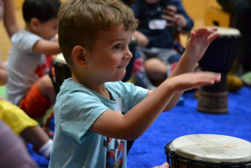A child playing a drum in a children's music workshop with other children seated on the ground.