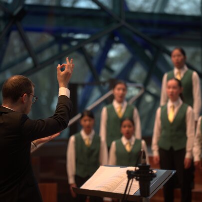 A conductor in a black suit with his arm in the air as he conducts the Australian Children's Choir in front of him.