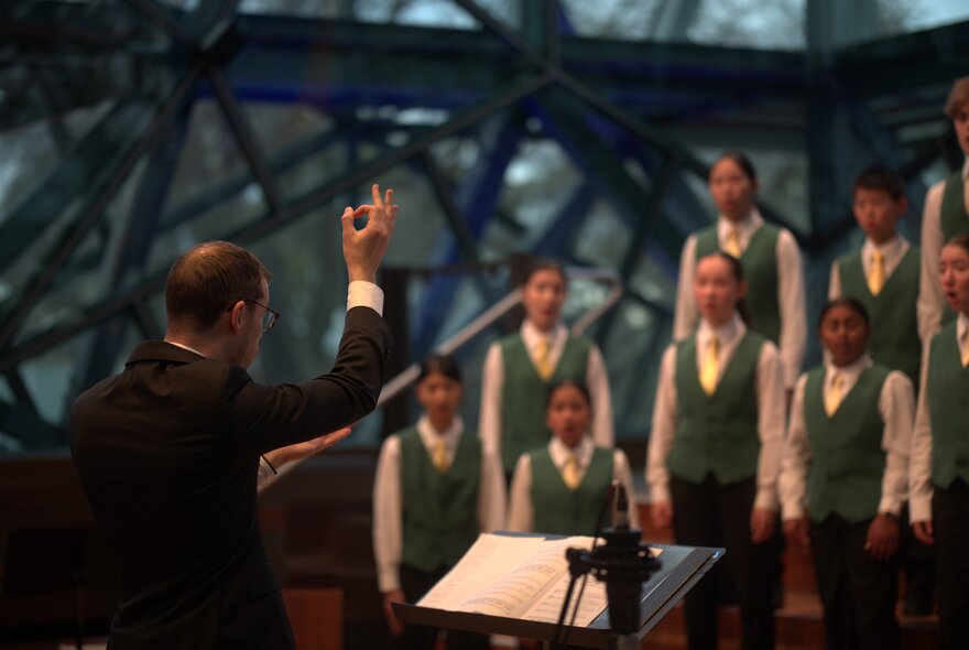 A conductor in a black suit with his arm in the air as he conducts the Australian Children's Choir in front of him.