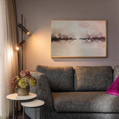A sofa in a hotel room with a magenta cushion and calming artwork, a lamp on in the corner.