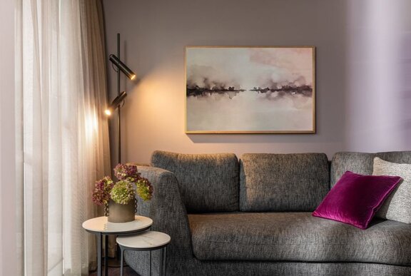 A sofa in a hotel room with a magenta cushion and calming artwork, a lamp on in the corner.