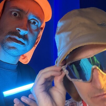 Radio and TV personalities Alex Dyson and Matt Okine, both wearing legionnaire's hats, one of them wearing sunglasses, and a neon glow coming from below. 