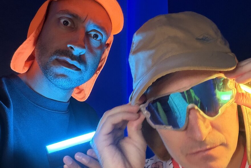 Radio and TV personalities Alex Dyson and Matt Okine, both wearing legionnaire's hats, one of them wearing sunglasses, and a neon glow coming from below. 